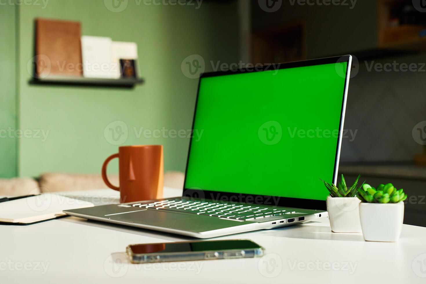 Modern home workspace featuring laptop with green screen, smartphone, coffee mug and small potted plants. Home office for freelancer with kitchen in background. Concept of productivity and remote work photo