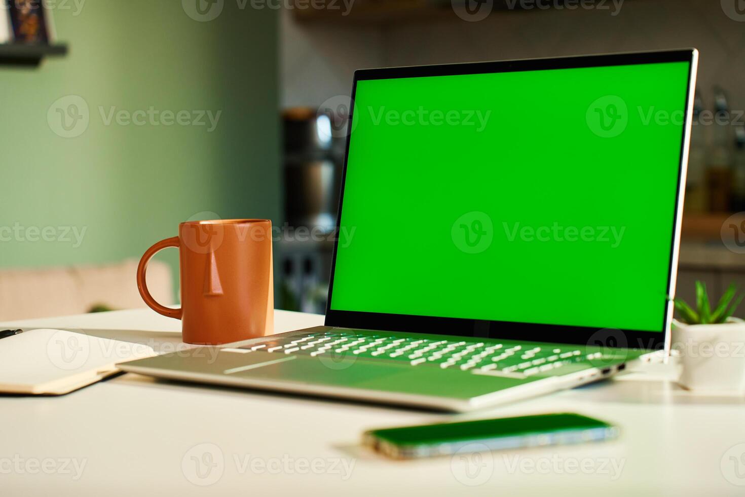 Modern home workspace featuring laptop with green screen, smartphone, coffee mug and small potted plants. Home office for freelancer with kitchen in background. Concept of productivity and remote work photo