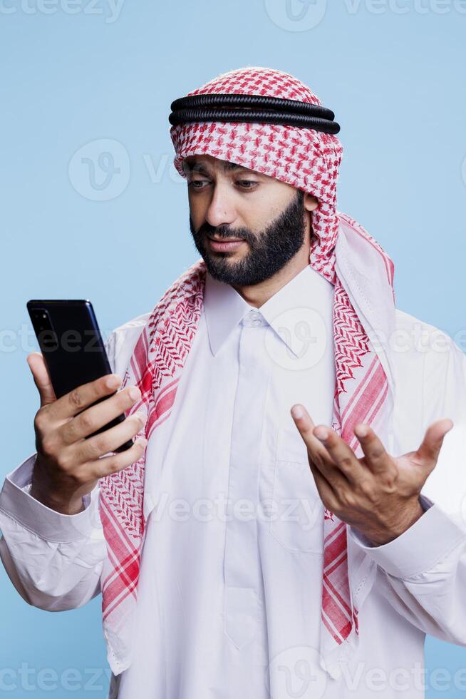 Arab man in traditional clothes engaging in virtual conversation on mobile phone. Person wearing muslim headscarf and thobe speaking in videocall and making gesture with hand photo