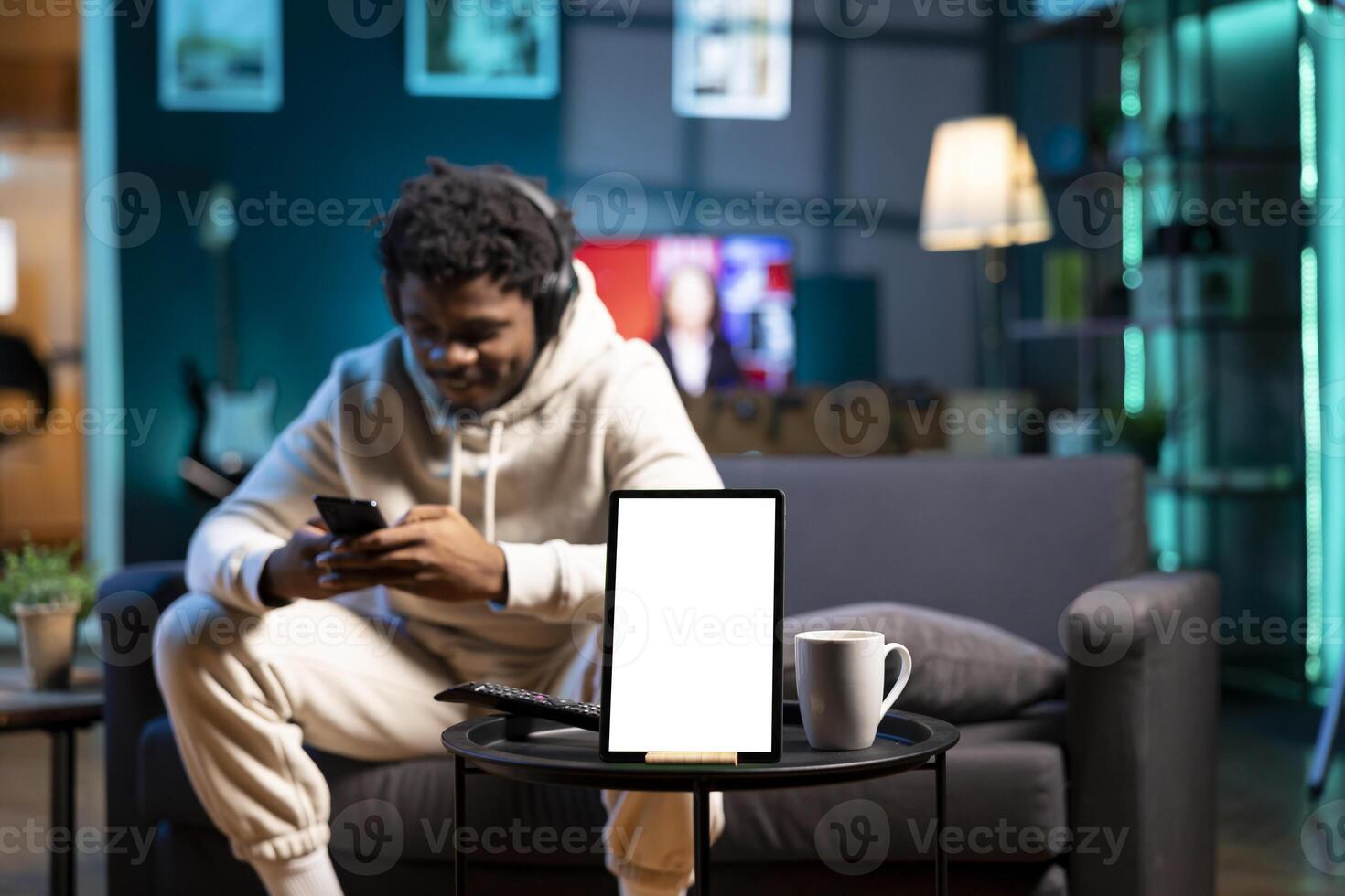 Focus on mockup tablet with man in blurry background sending messages on phone and listening music. Close up of isolated screen device in front of person wearing headphones, texting friends photo