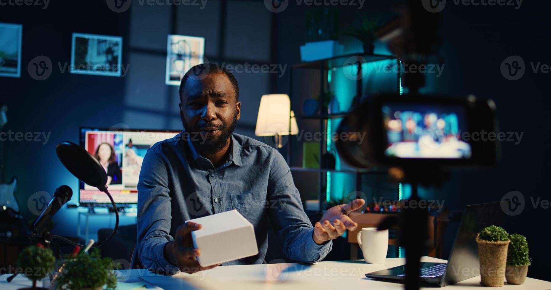African american tech content creator filming packaged product unboxing in RGB lights personal home studio. Influencer showing sponsoring branded merchandise, urging subscribers to purchase photo