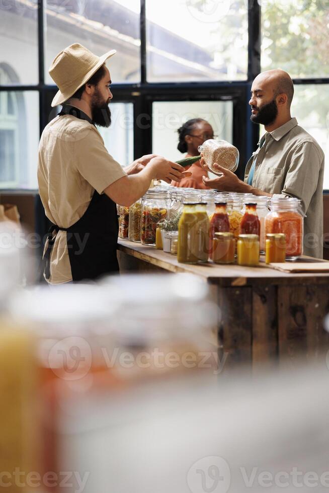 Male storekeeper in black apron hands over green vegetable to customer holding jar filled with organic food item. Salesman offering advice to client shopping for pantry staples at sustainable shop. photo