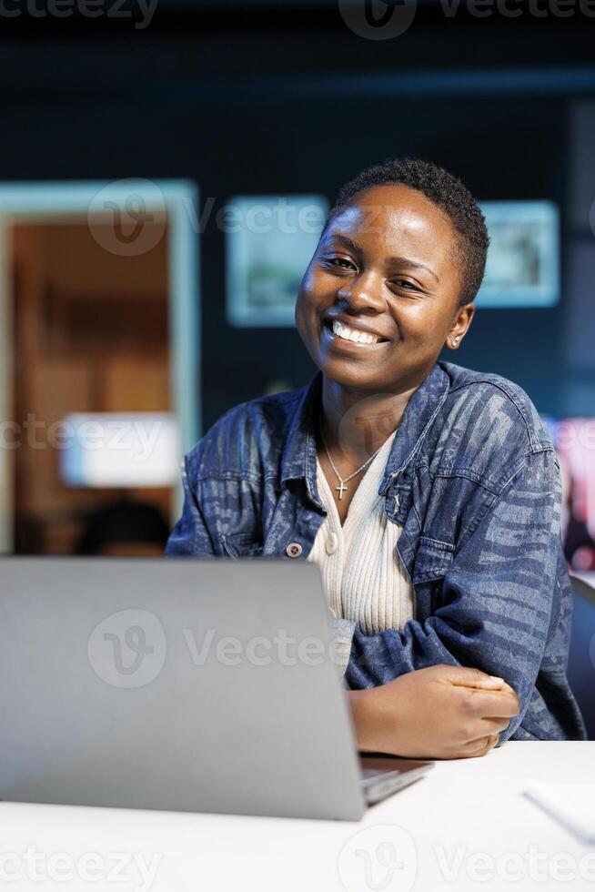 Portrait of African American influencer seated in front of a portable computer, grinning sincerely. Cheerful black woman sitting, looking at camera with her arms crossed, close to her laptop. photo