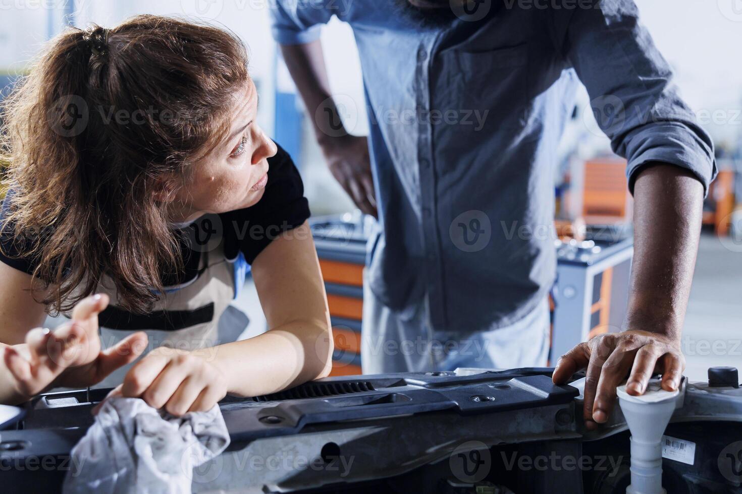 Diligent serviceman in garage finishing refurbishing car, looking underneath vehicle hood to remove remaining oil leaks. Employee does yearly brakes system cleaning on customer automobile photo