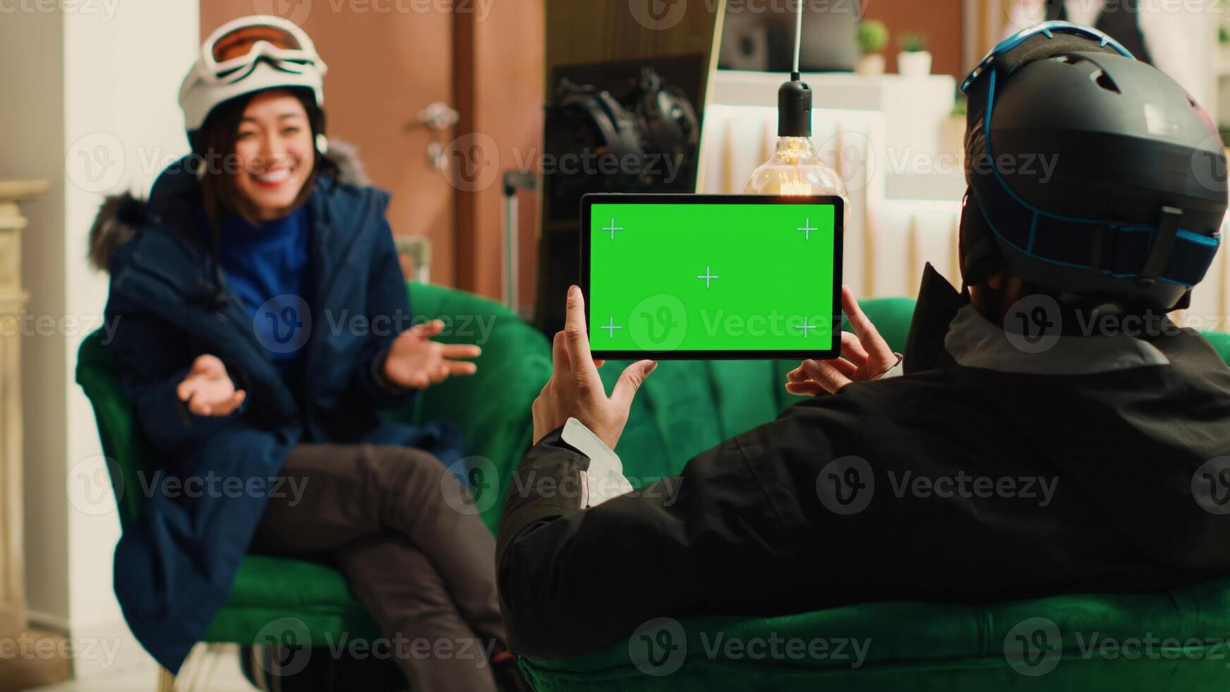 Young man uses tablet with greenscreen display in ski resort lounge area, holding digital gadget displaying blank chroma key mockup template. Couple talking about extreme winter sports. photo