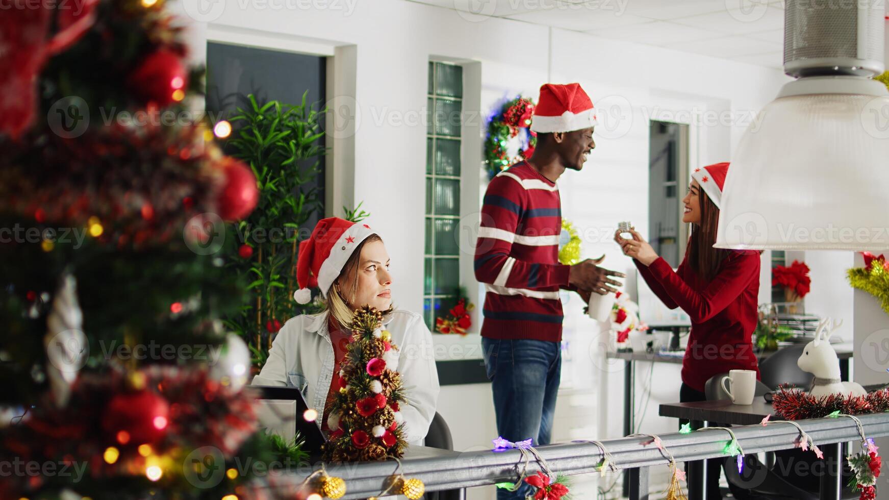 Focused employee in Christmas decorated office working on overdue project while carefree coworkers enjoy coffee break together looking at decorations. Stressed worker in festive ornate workspace photo