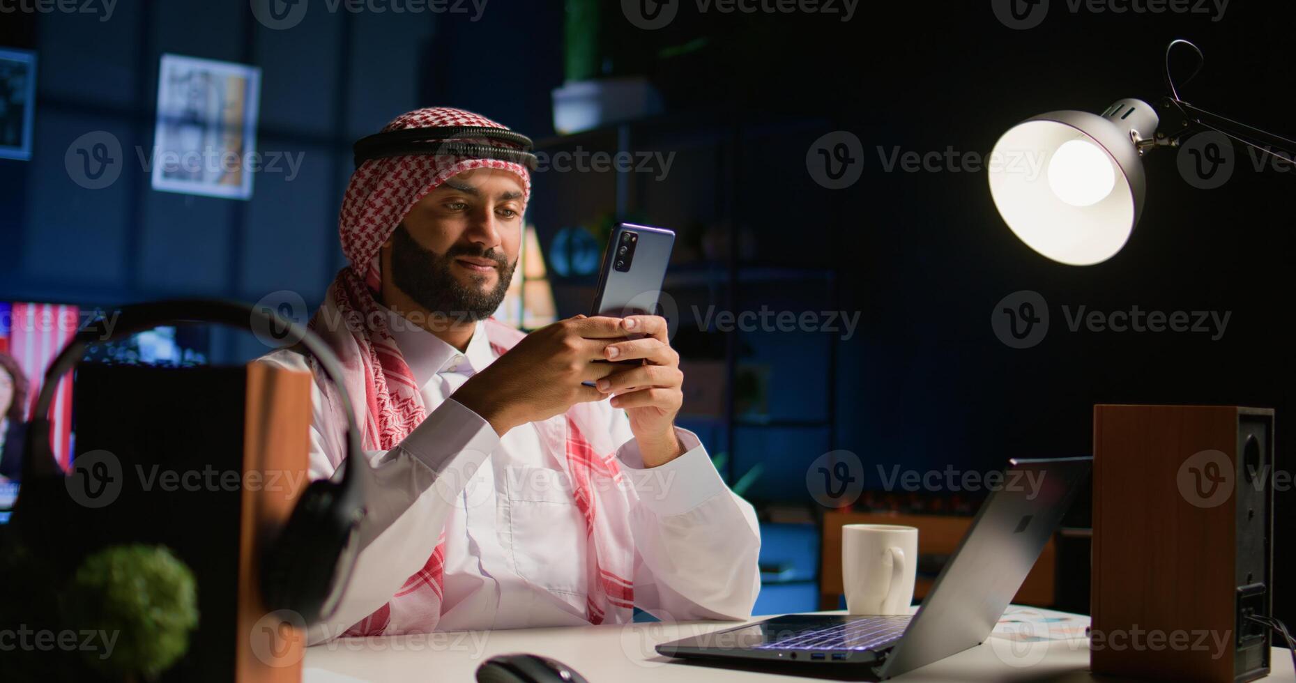 Cheerful arab man at home in living room writing messages on smartphone. Middle Eastern person holding cellphone, enjoying relaxing leisure time talking with friends over internet texting app photo