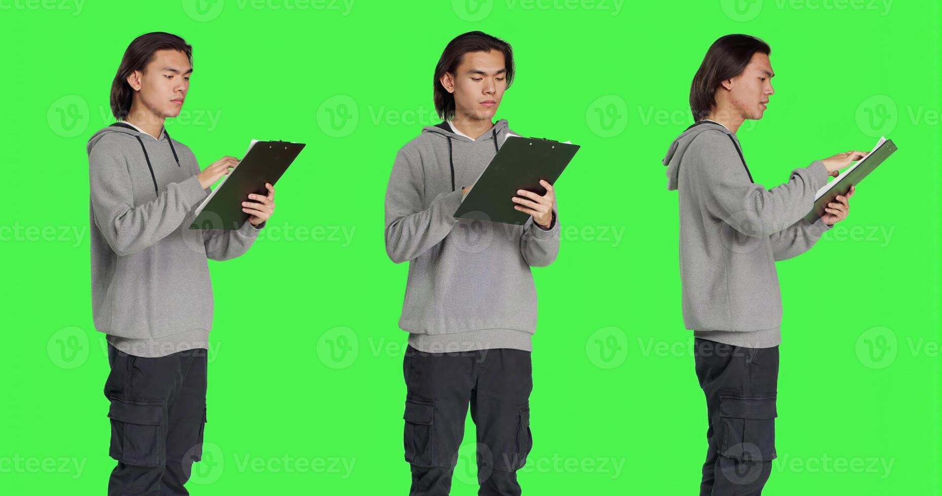 Asian guy reads information on files, thinking about annotations on clipboard papers or taking notes for completion. Young man writing on forms, standing against greenscreen backdrop. photo