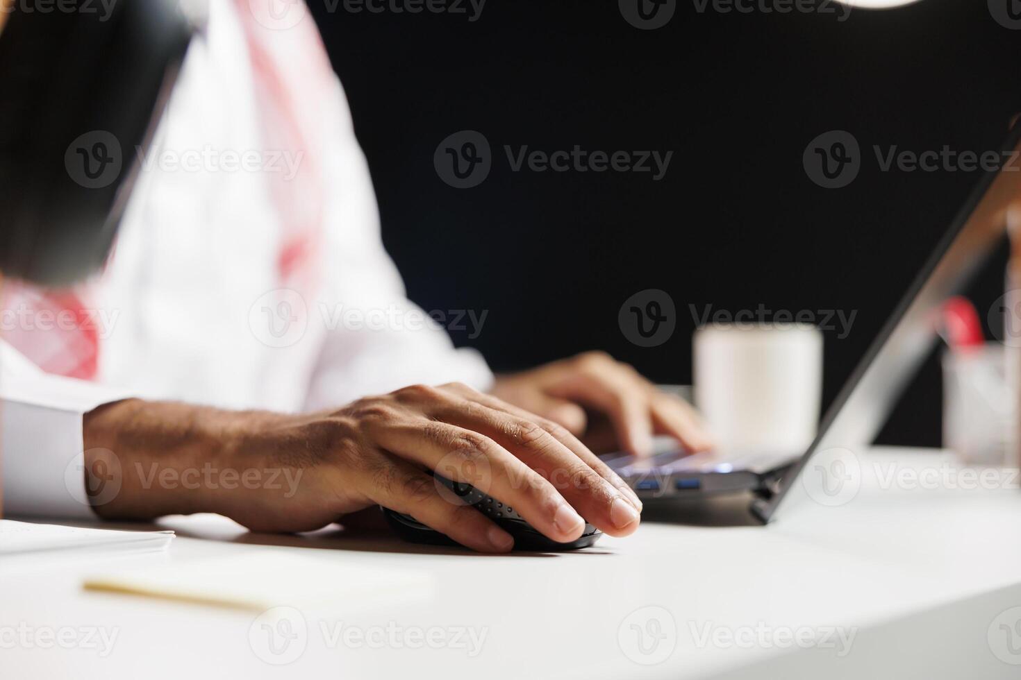 Close-up view of a pair of male hands using a wireless mouse and laptop, doing online research. Detailed shot of a person with Arabian clothing making use of the minicomputer on the table. photo