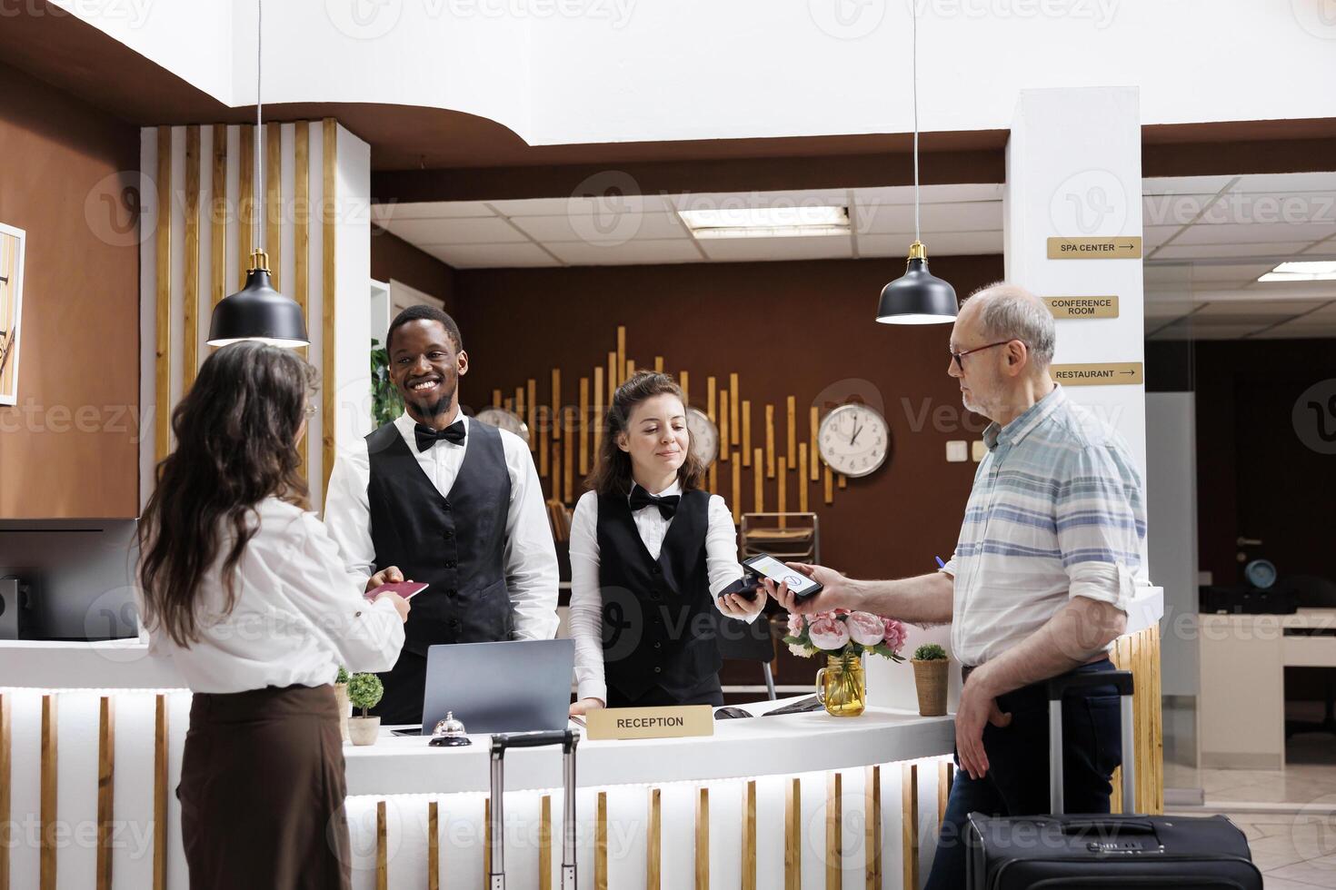 Elderly couple receives assistance from friendly employees at hotel reception counter. Old man using mobile phone to pay for check-in while senior woman gives passport for verification of reservation. photo