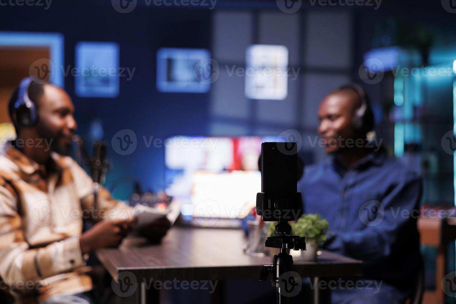 The podcast host uses smartphone on a tripod to record guests giving fans fascinating stories from living room. Invited to the home studio, male celebrity shares facts with interviewer. photo
