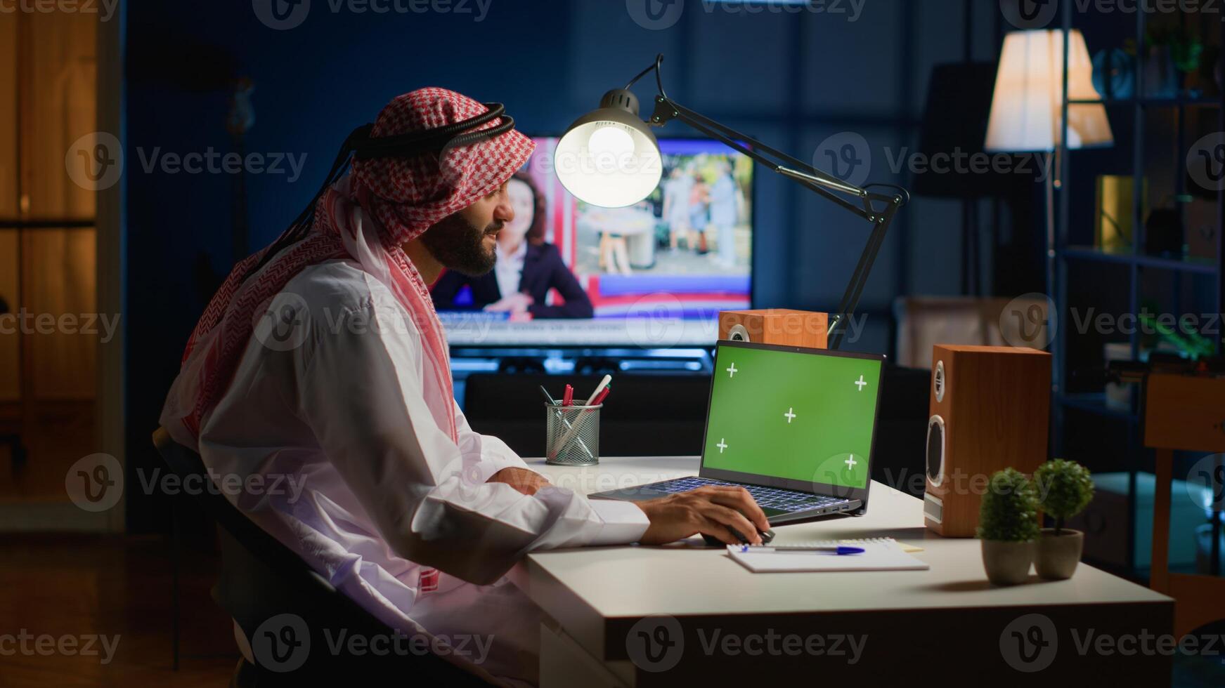 Arab remote worker inputting data on mockup laptop while working from home. Self employed freelancer performing tasks on isolated screen device in apartment office with TV as background noise photo