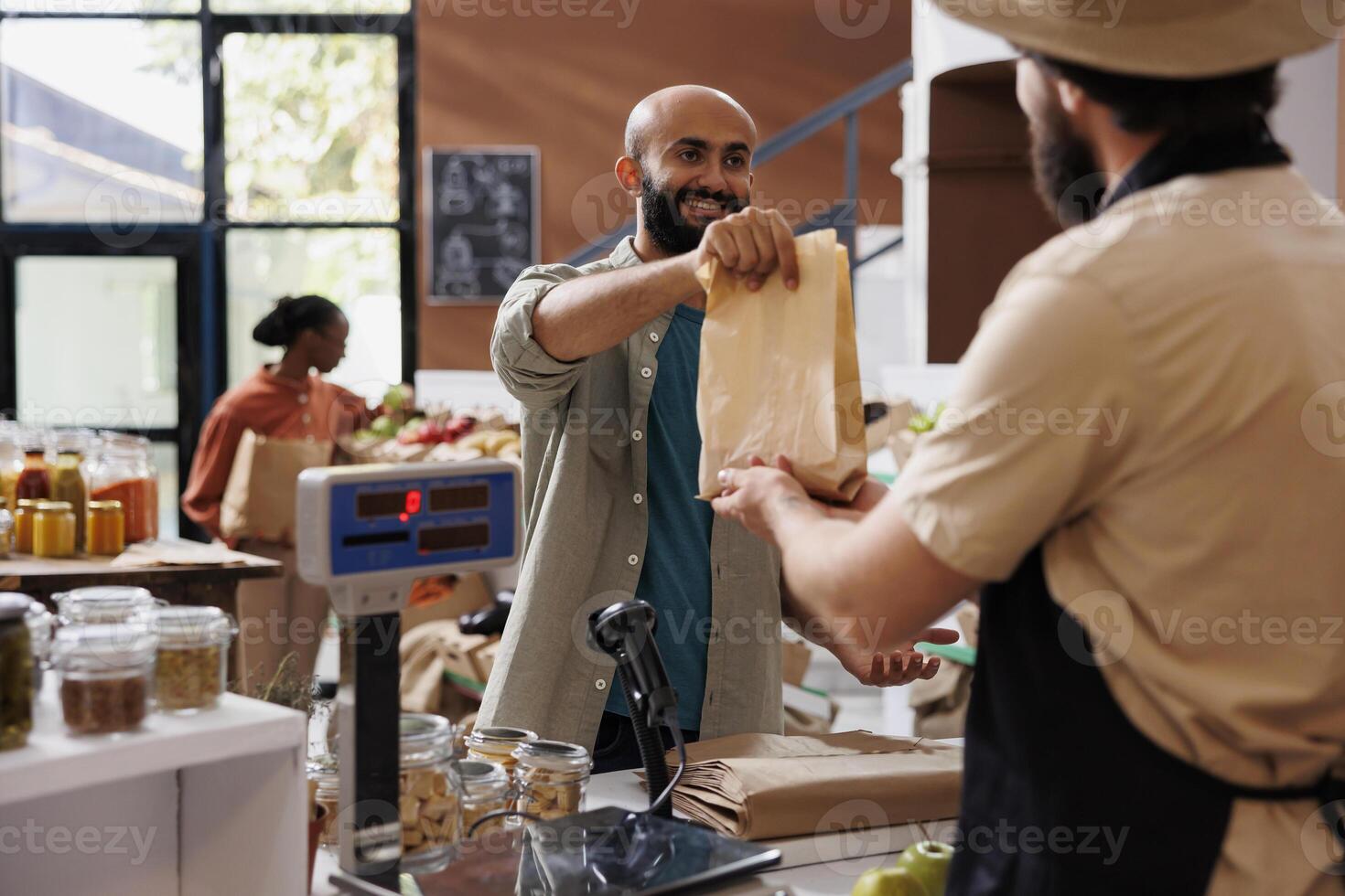 Smiling customer choosing organic products to buy and approaching vendor at counter to pay for bag of homegrown produce. Buyer doing grocery shopping at local zero waste eco store. photo