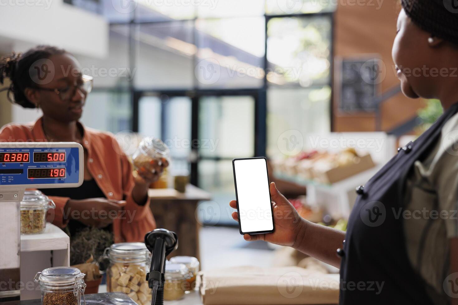 Woman in zero waste shop using mockup mobile device to analyze products. Vegan customer checking food item suitable for his diet while female vendor holds smartphone with isoalted white screen. photo