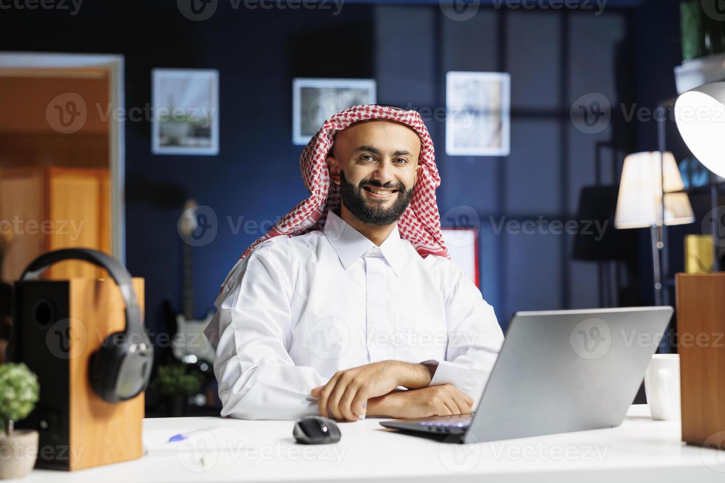 Profile of smiling male Arab entrepreneur sitting at a desk with his personal computer nearby. Portrait shot of a cheerful Muslim man with a laptop on the table, utilizing modern technology. photo
