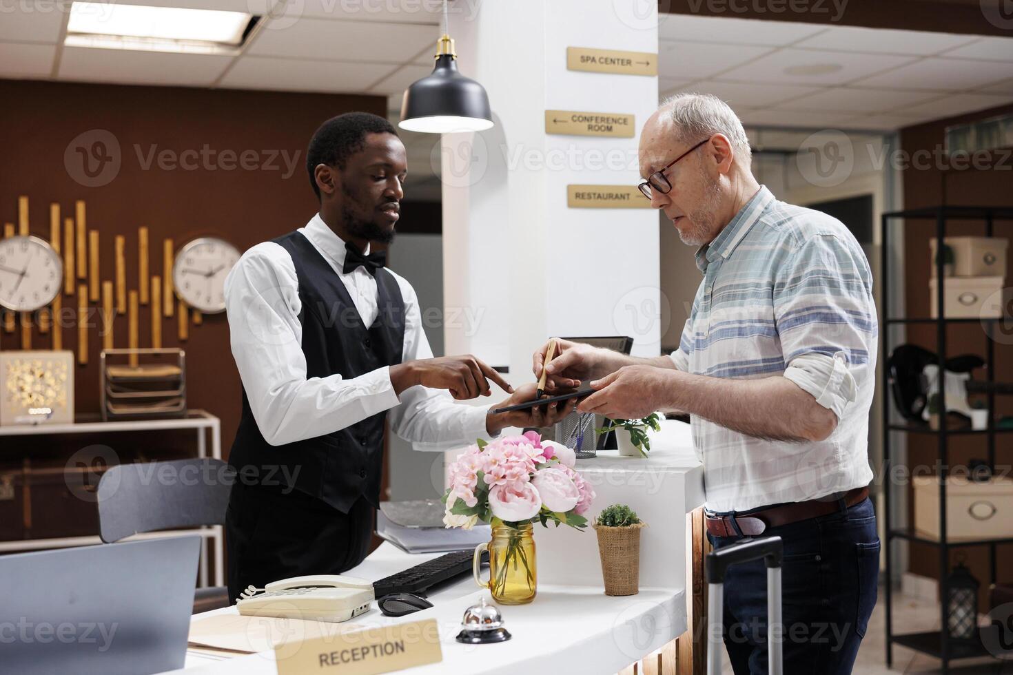 Mature old man standing at hotel reception counter, where polite receptionist aids with reservation procedure on digital tablet. Elderly male client signs an online check-in form on smart device. photo