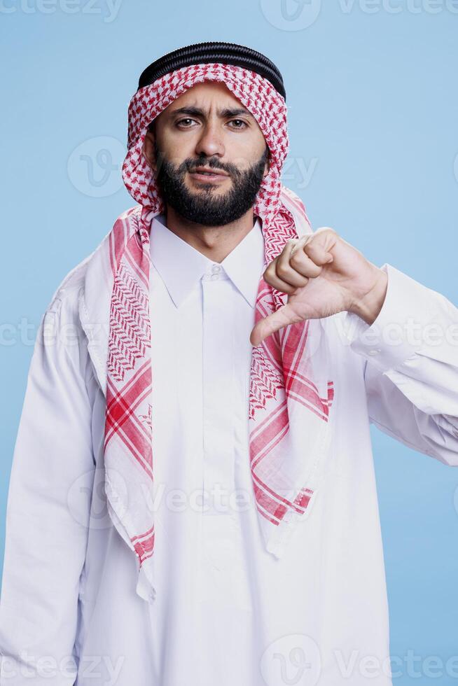 Arab man in traditional robe and headdress showing thumb down studio portrait. Confident muslim person wearing white thobe showcasing disapproval gesture and looking at camera photo