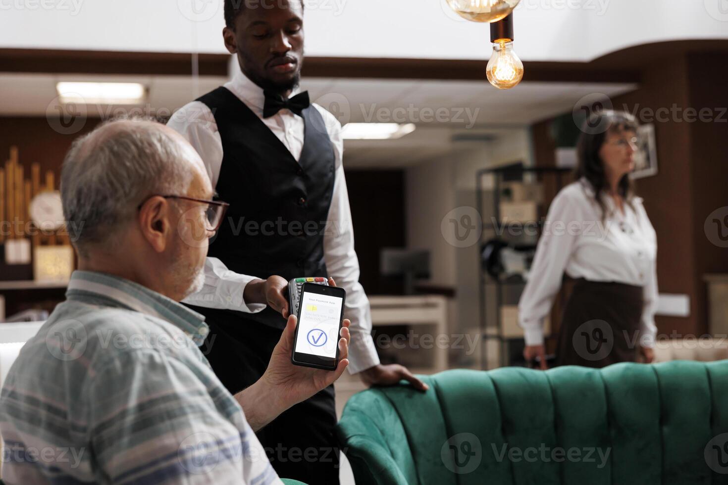 Retired senior male traveler in cozy lounge area paying with smartphone on nfc terminal. African american waiter in hotel lobby assisting elderly man with cashless payment using mobile device. photo