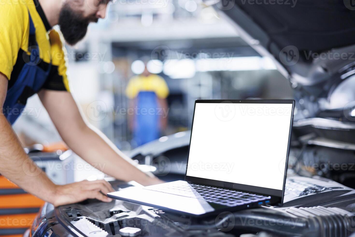 Mockup laptop on car with hood open while mechanic in blurry background replaces fuel tank. Isolated screen device next to meticulous garage worker repairing client vehicle photo