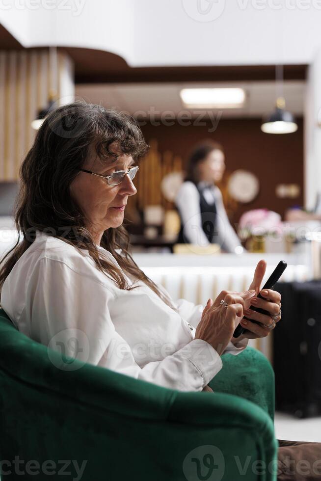 Detailed view of elderly female client seated in hotel foyer, using her cell phone to browse the internet. Retired old woman checking in online using mobile device. Close-up, side-view. photo
