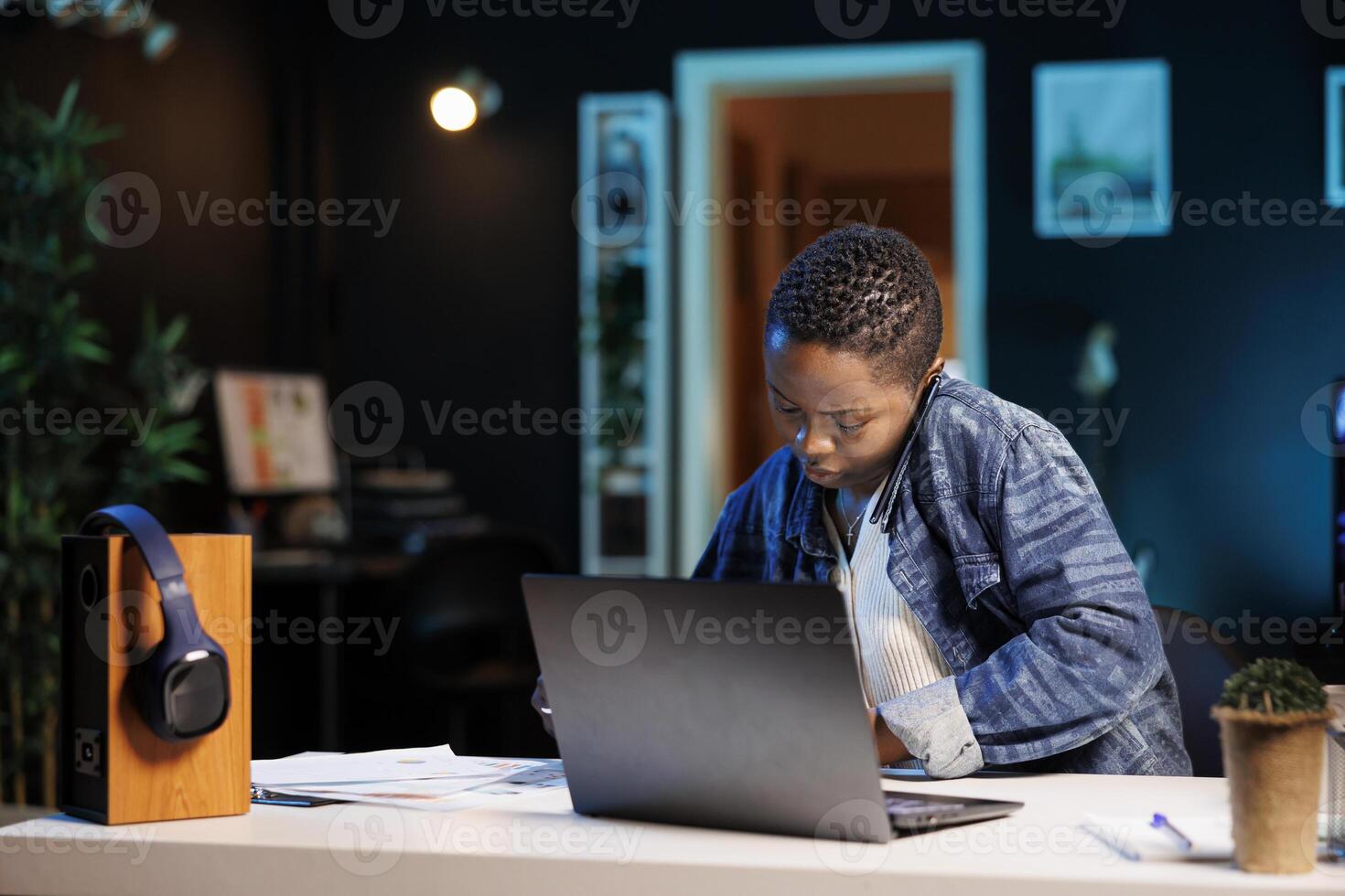 Black woman working from home at desk, using digital laptop and smartphone. African American freelancer uses mobile device for communication and a wireless computer for typing during phone call. photo