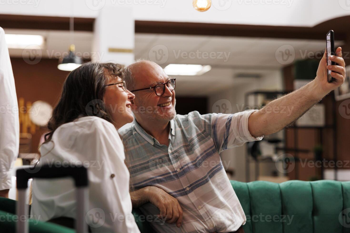 Senior old man holding cell phone for selfie with wife in exclusive hotel lobby. Retired elderly couple with suitcase, sitting in lounge area with mobile device in hand for a call. photo