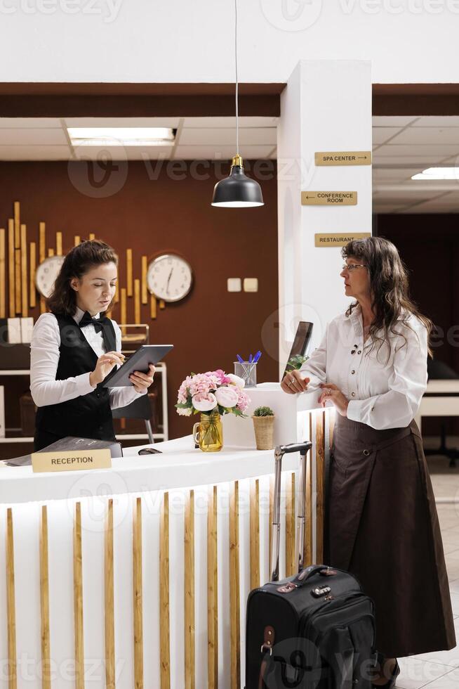 Elderly lady stands at hotel reception, where professional concierge assists with reservation. Senior woman completes check-in process with digital document on tablet. Suitcase is placed near counter. photo