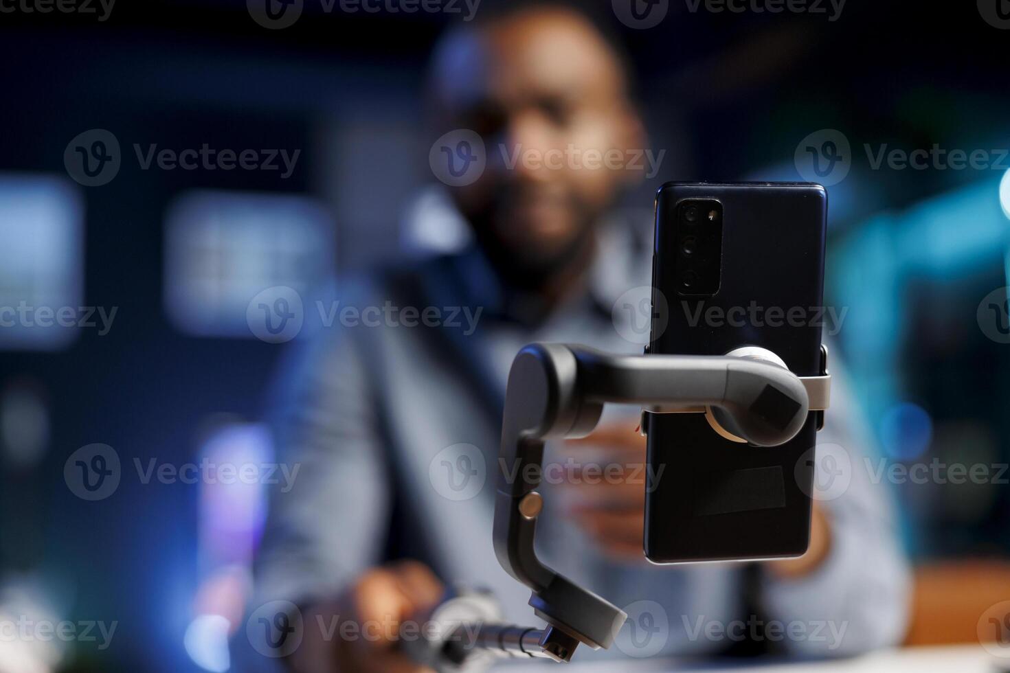 Enthusiastic internet show host in blurry background hitting the record button on cellphone. Focus on mobile phone attached to selfie stick used by content creator to record footage for fans photo