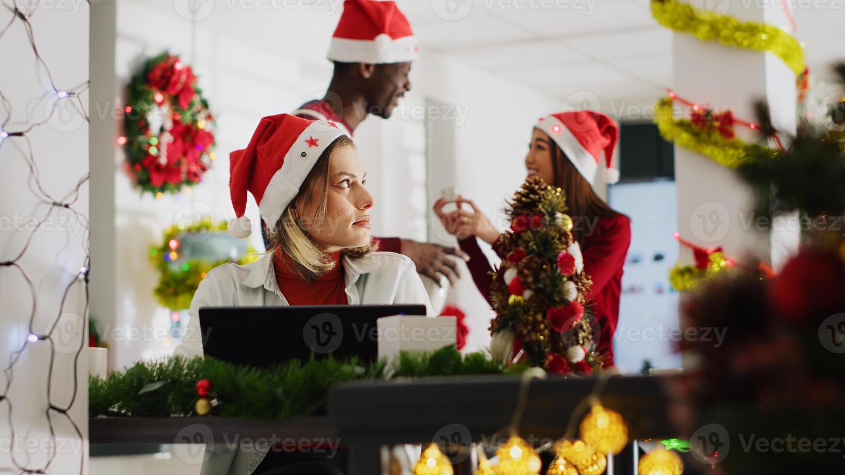 Woman filling paperwork in Christmas decorated office, perturbed by disturbing staff members discussing in background. Employee unable to concentrate on tasks while colleagues talk loudly photo