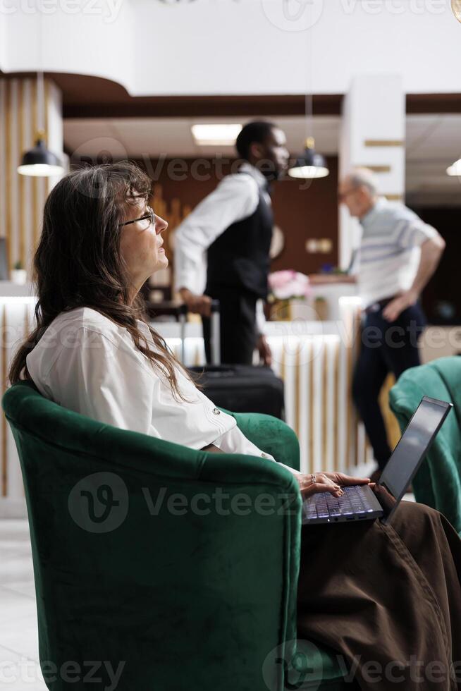 Side-view shot of elderly female customer sitting in hotel lounge area, browsing the internet on her laptop. Close-up of retired senior woman using her personal computer for online check-in. photo