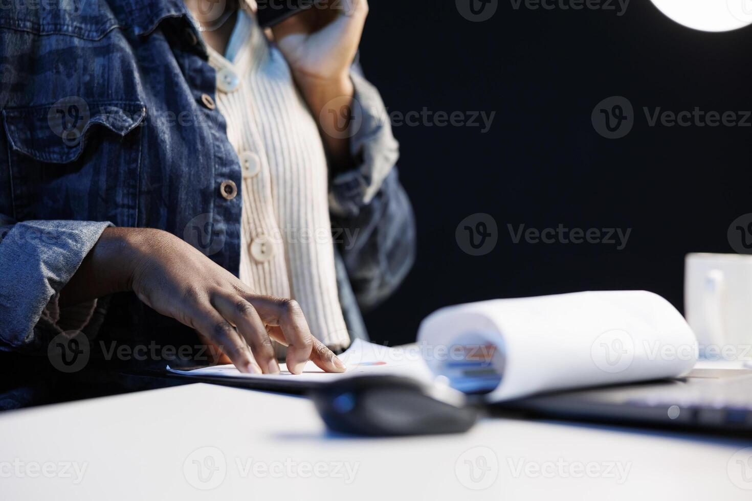 Close-up of black individual using smartphone to talk while studying documents at a desk. African American freelancer with her hand on a clipboard as she checks and examines papers, working from home. photo
