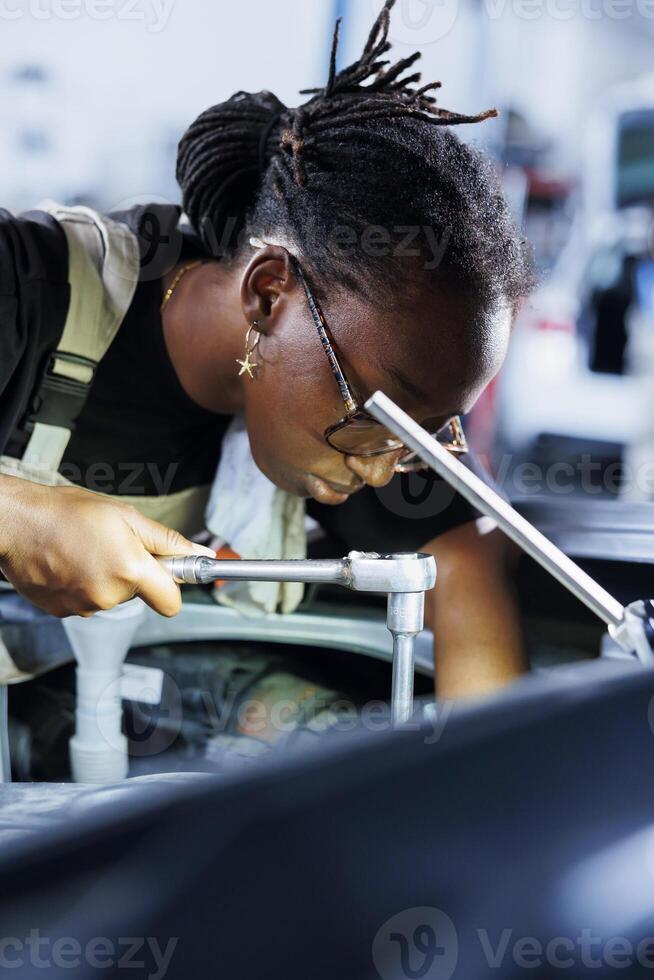 BIPOC technician does client car maintenance in auto repair shop using lamp light after vehicle shut down unexpectedly. Trained expert in garage mending customer damaged automobile fuel tank photo