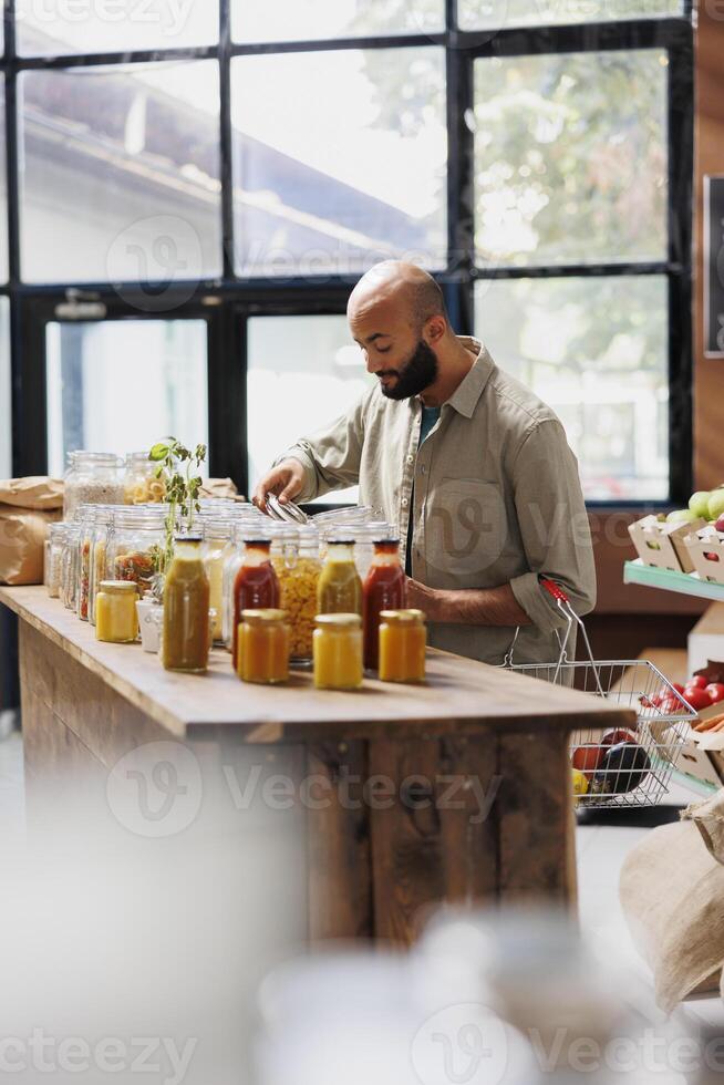 Young man carrying shopping basket while looking at jars of different fresh food products. Male consumer searching for sustainable items with plastic free packaging at local convenience store. photo