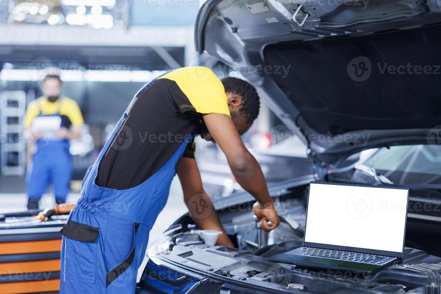 Mockup laptop on car with hood open while repairman in blurry background replaces motor. Isolated screen device next to licensed garage specialist fixing customer automobile photo