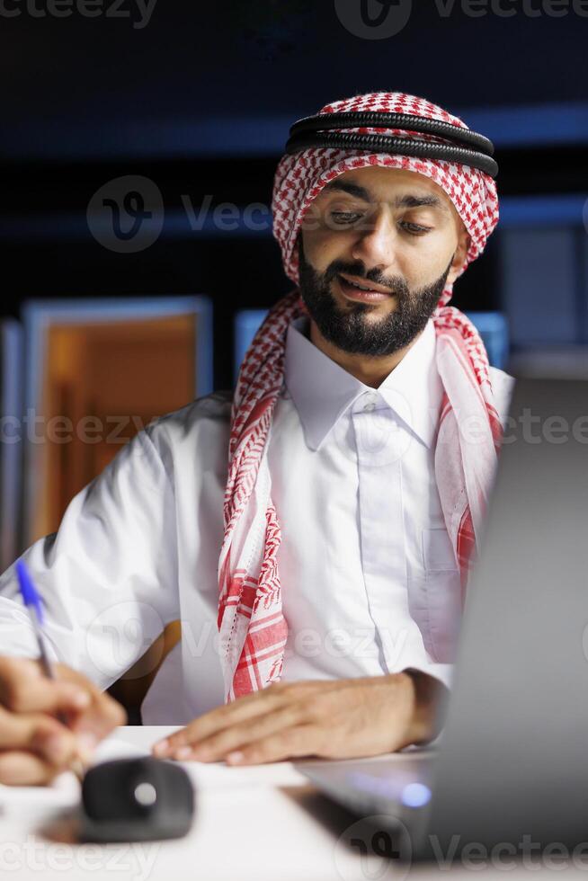 Detailed view of Arab man working efficiently at the workplace by surfing the internet and writing notes on a book. Close-up of Muslim guy doing a study with his notepad and personal computer. photo