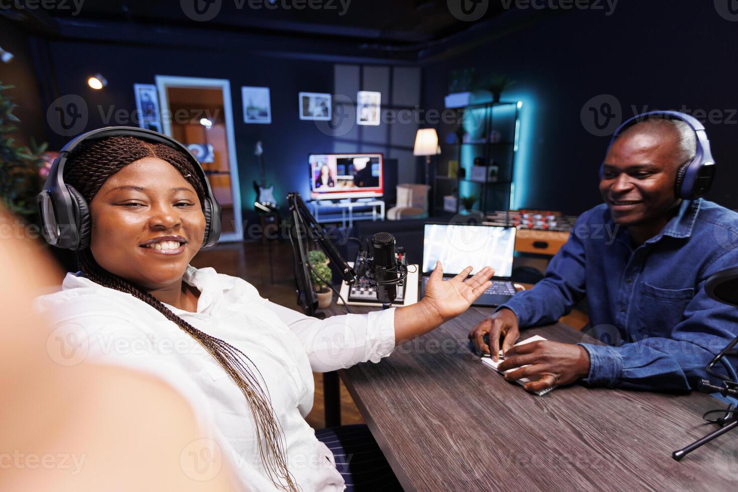 African lady filming podcast with guest telling listeners an engaging narrative using selfie camera on cell phone. Female interviewer introducing male influencer to online audience using mobile device photo
