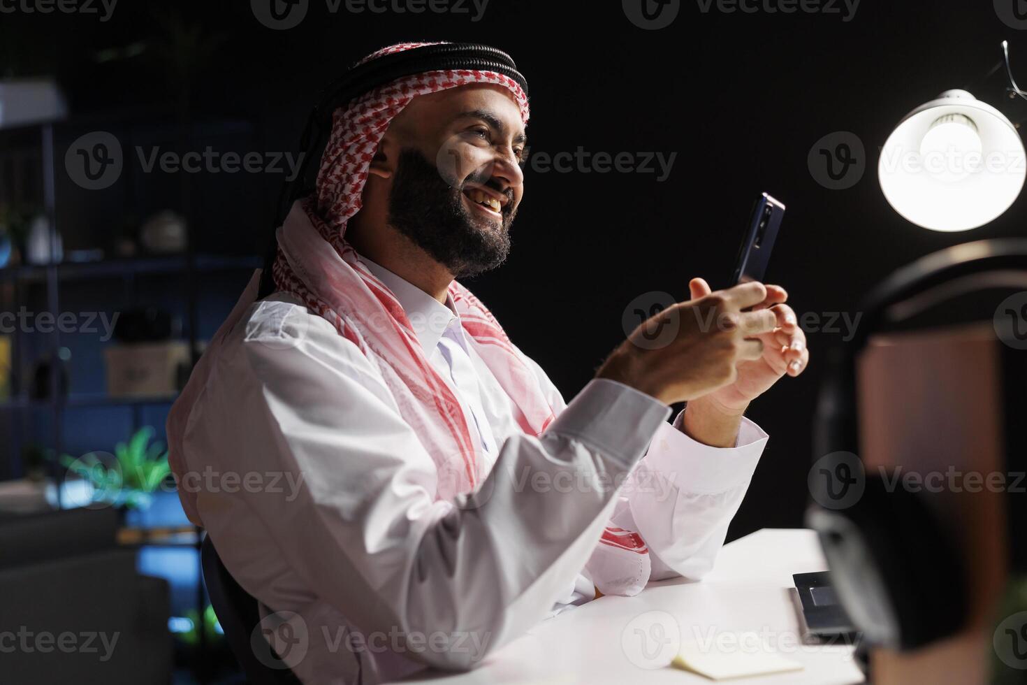 Side-view of smiling Muslim man in professional attire works at a modern desk. Using a smartphone, he browses the internet, representing the digital age and wireless technology. photo