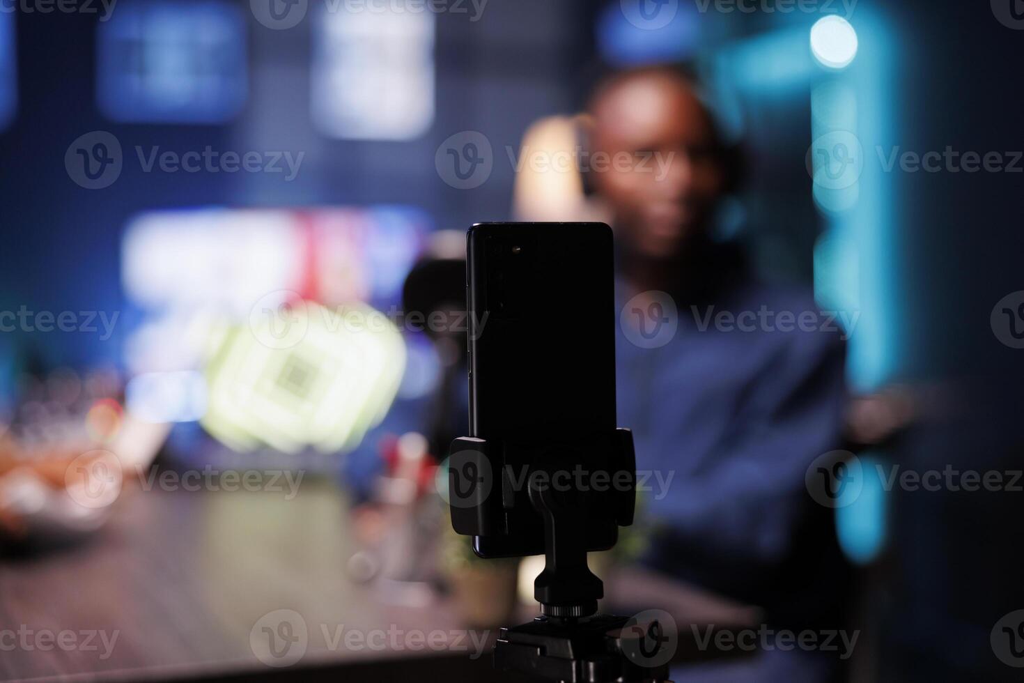 Focus on smartphone on tripod recording vlogger show host in blurry background interviewing guest in neon lights studio. Detailed view of mobile device being used to film content for male influencer. photo