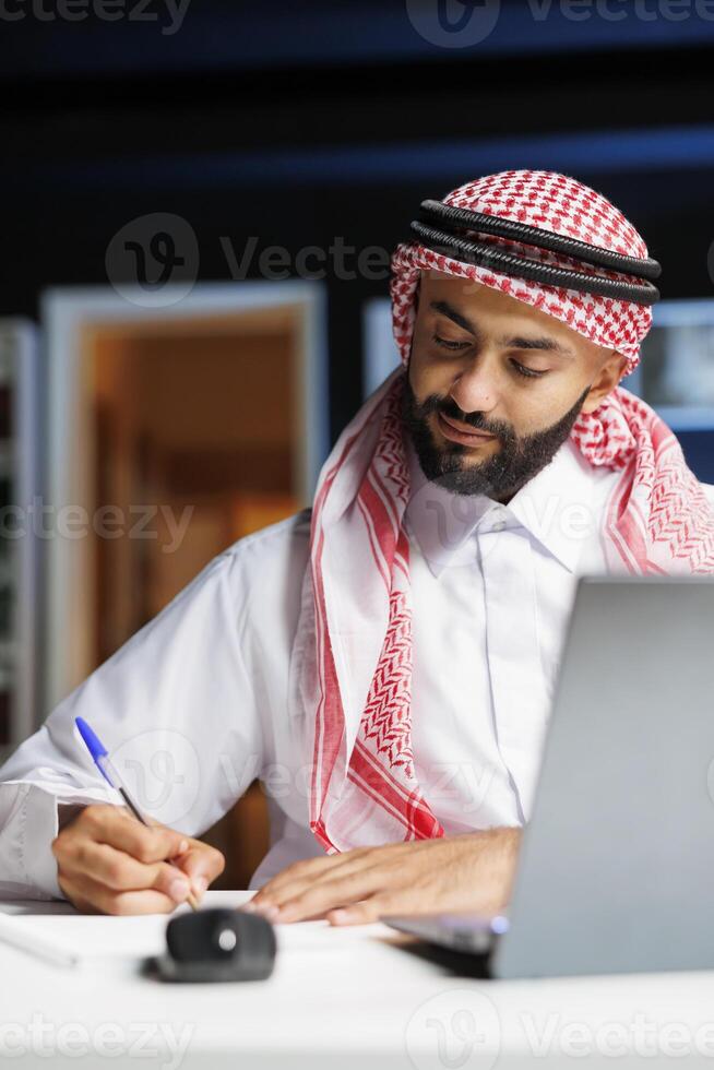 A focused Middle Eastern man working at a table with a notebook is shown in detail, dressed traditionally. A close-up of a Muslim man writing out research at his desk at home. photo