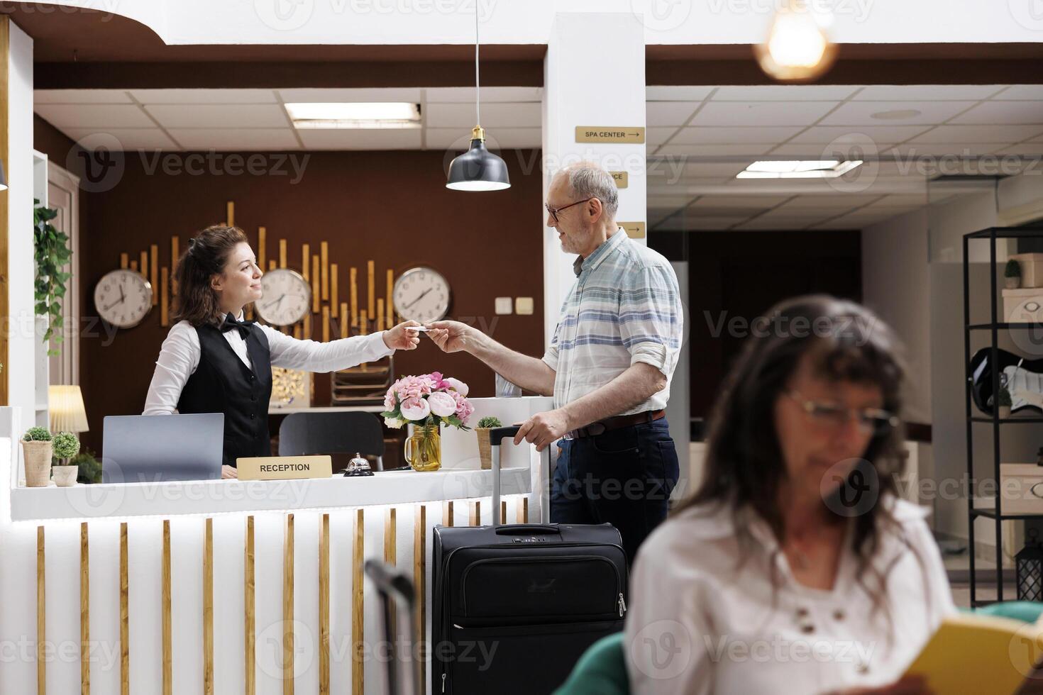 Caucasian old man takes room key card from female concierge after doing registration at front desk of hotel lobby. Elderly male tourist finishes check-in process at luxury registration counter. photo