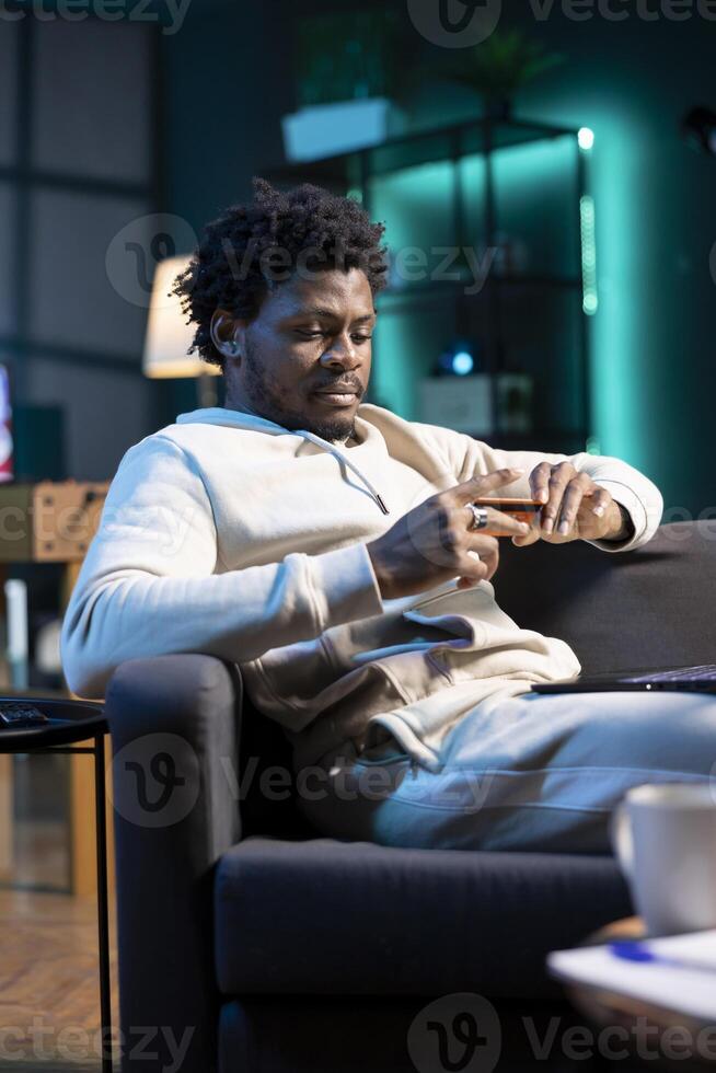 Man doing internet purchases after receiving pay rise at work, typing credit card information on computer. BIPOC person adding payment method on website late at night in neon lit room photo