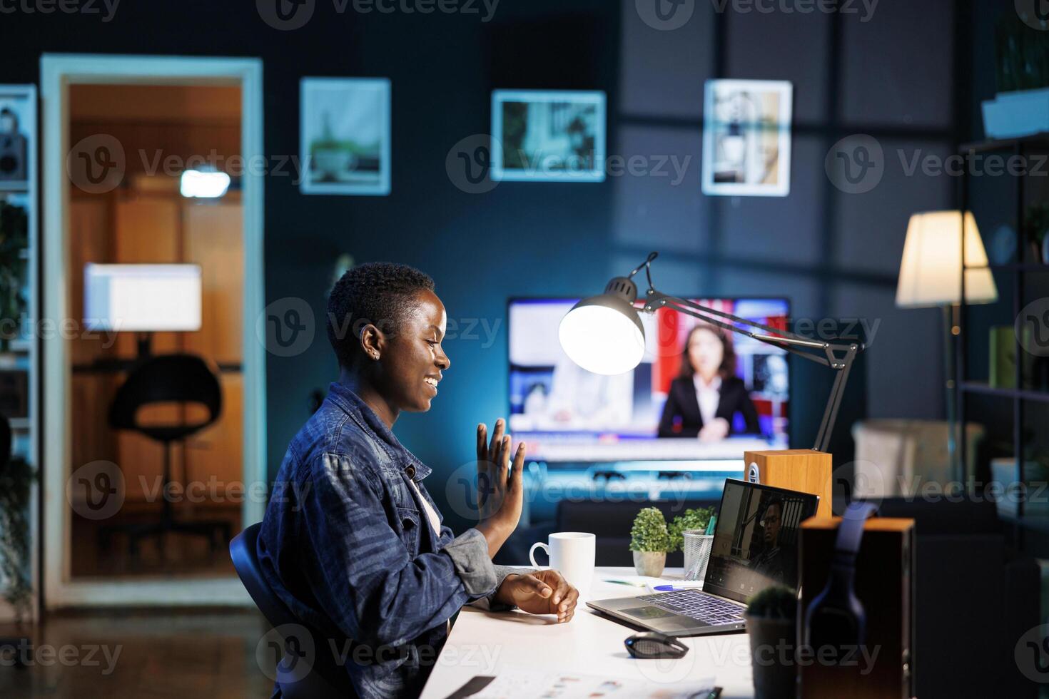 African american blogger waving at her personal computer while seated at a desk having a call. Black woman uses digital laptop to chat online and research while immersed in digital technology. photo