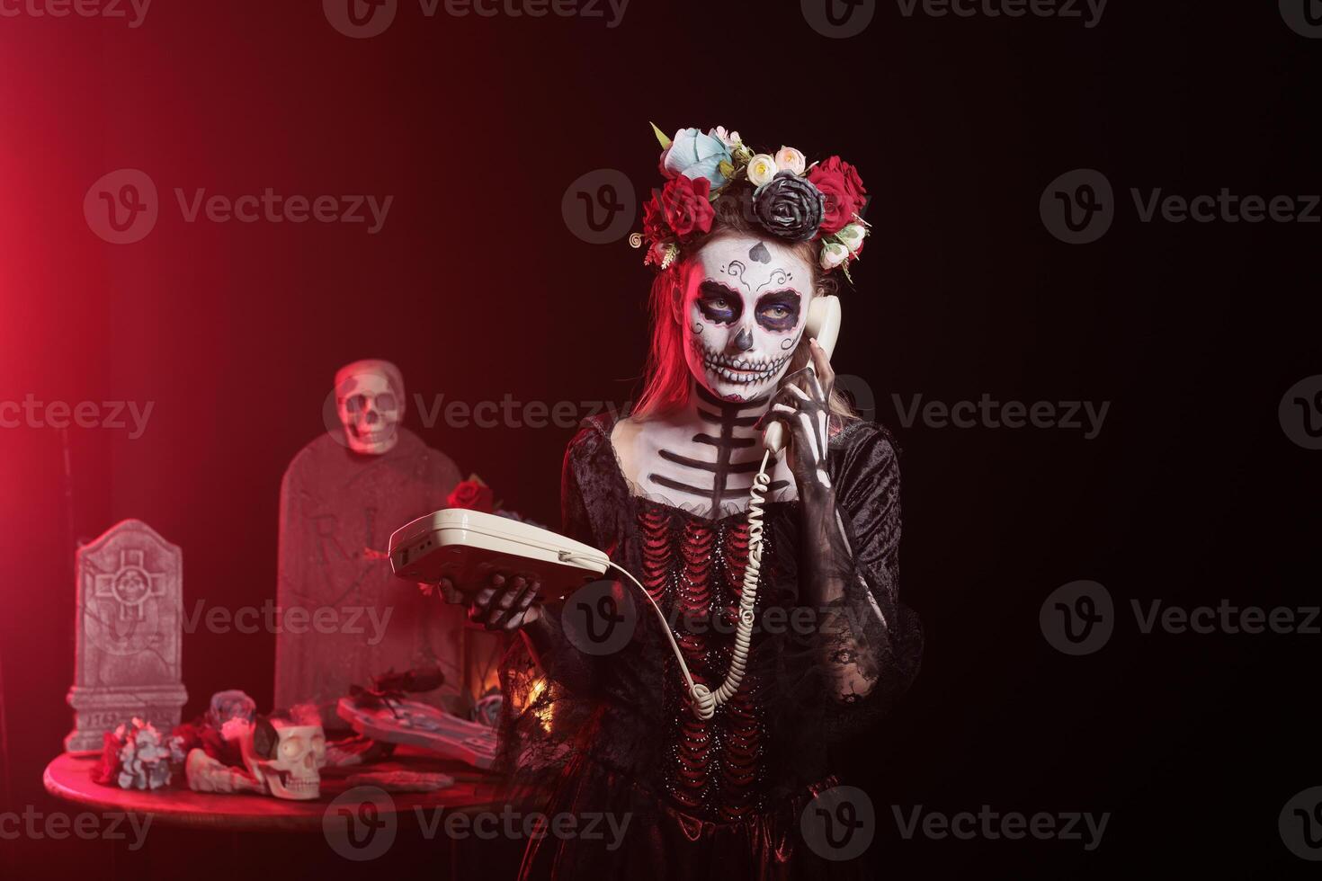 Woman using landline phone to answer call in graveyard, celebrating Day of the Dead. Mysterious lady talking on office cord telephone surrounded by frightening funeral items during Halloween event photo