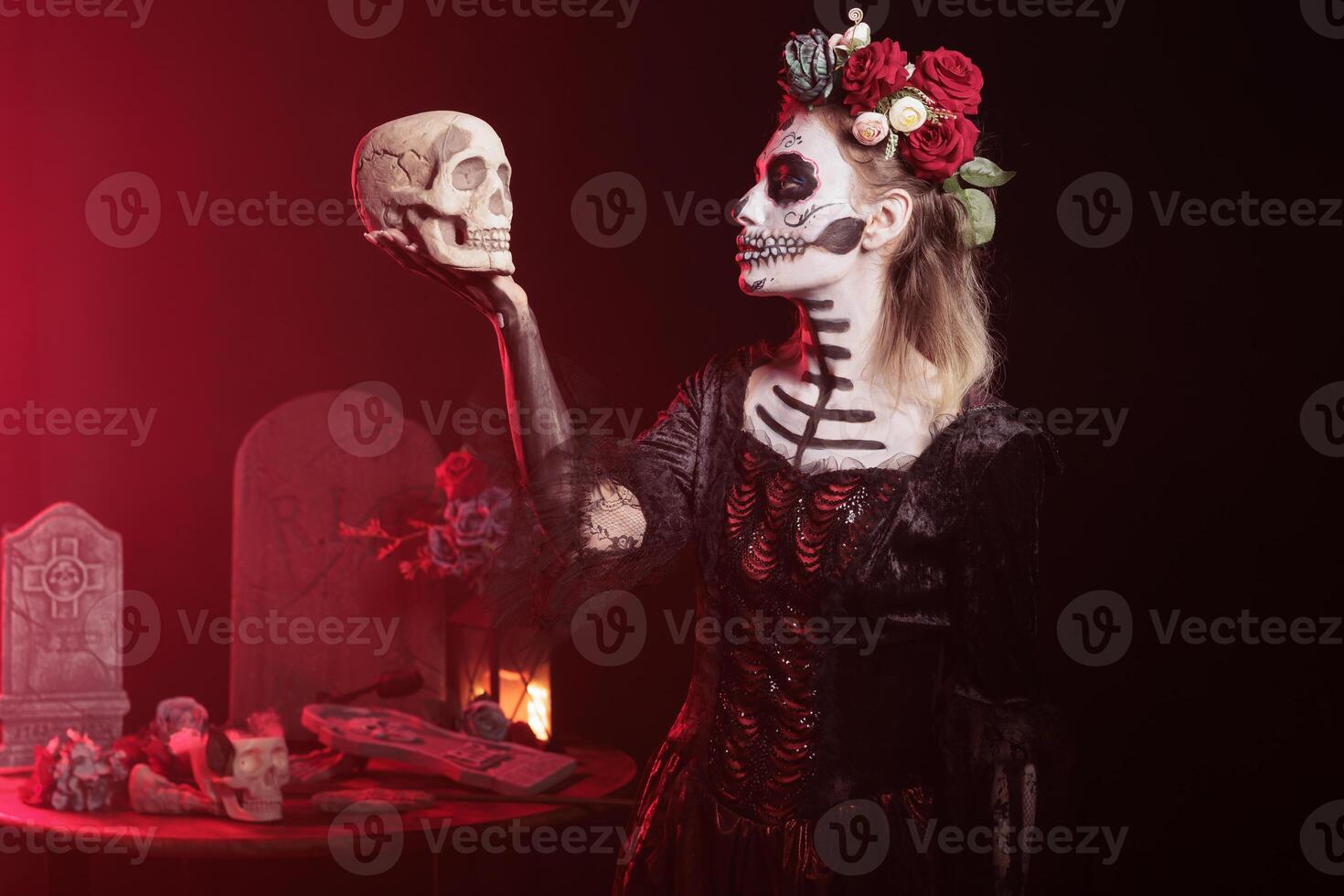 Woman posing with skull in hand, acting Hamlet scene while wearing skeleton makeup. Macabre lady dressed as La Catrina character on day of the dead event in studio background with red light photo