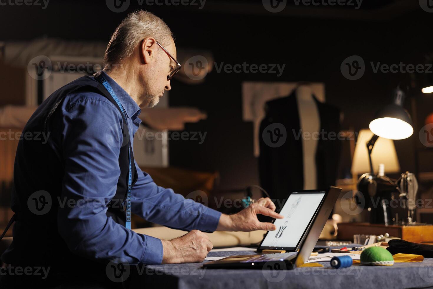 Elderly skilled dressmaker zooming on tablet, looking at sketch drawings of sartorial custom made outfits. Fashion designer working on customer comission in professional atelier shop workspace photo