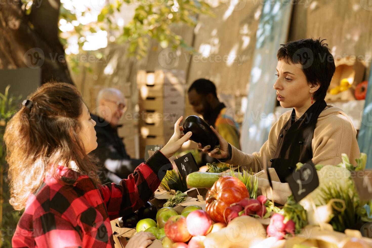 Vendor woman talking to female customer and selling fresh locally grown produce at farmers market stand. Seller helping client to choose veggies, explaining health benefits of organic food. photo
