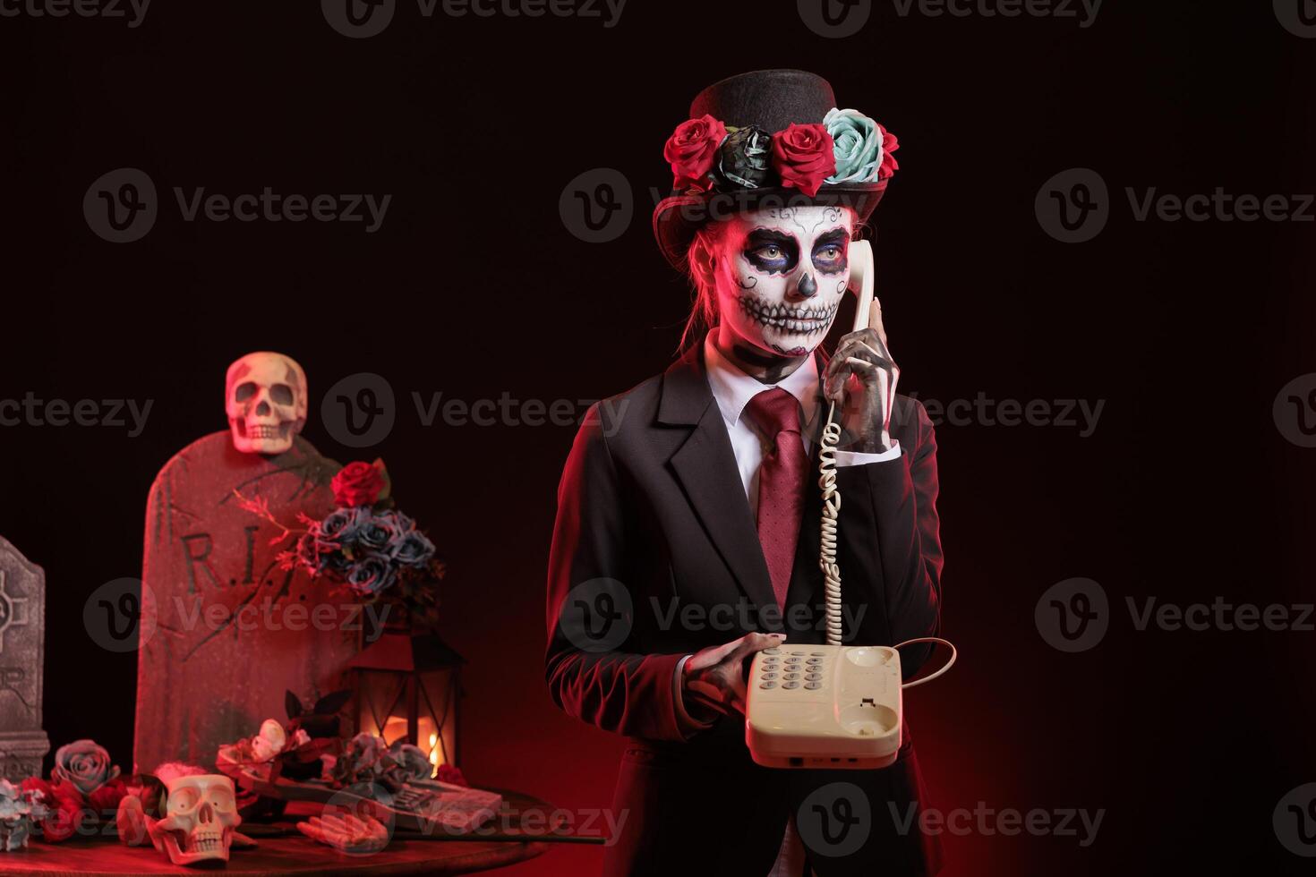 Woman wearing skeleton paint uses landline phone to answer call in graveyard, celebrating Day of the Dead. Lady with corpse makeup talks on cord telephone surrounded by funeral items during Halloween photo