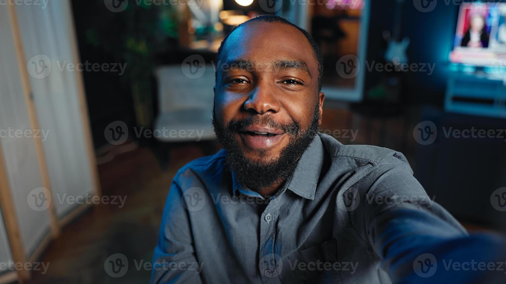 Smiling influencer using mobile phone to do live broadcast for subscribers, showing his home studio. Internet star films himself for viewership using POV camera, engaging with fans watching photo