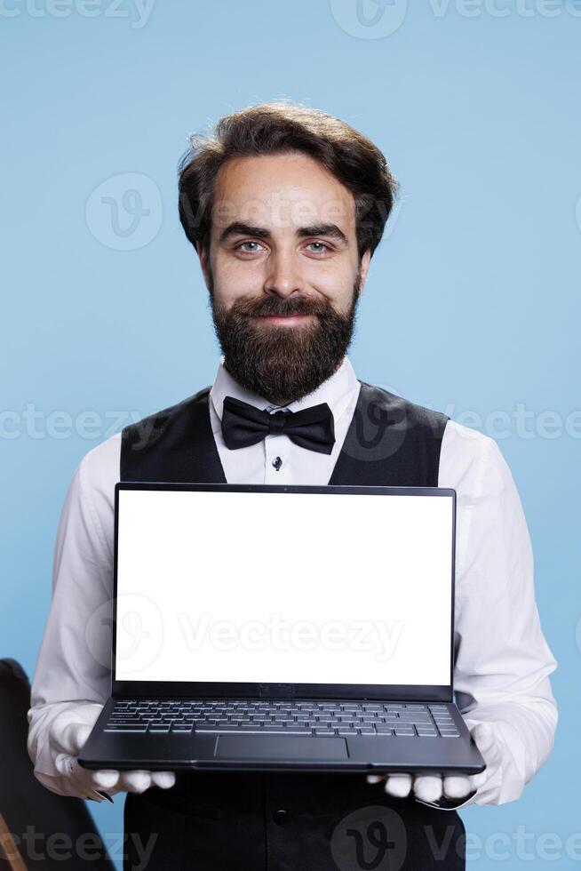 Young bellboy holds white display on laptop in studio while he wears formal suit and tie next to luggage. Classy doorkeeper working at luxurious hotel and shows blank screen template. photo