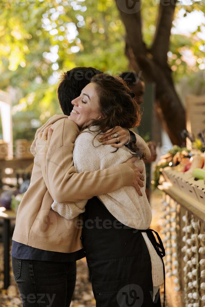 Smiling female individuals hugging each other at eco friendly farmers market, happy vendor enjoys visit from favorite customer. Caucasian merchant feeling positive about small business clients. photo